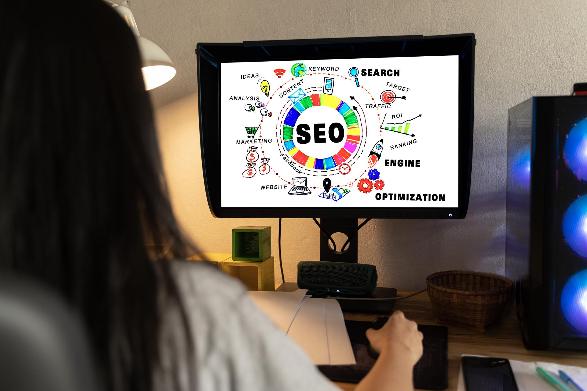 A girl sitting at a desk with a laptop showing an SEO diagram, surrounded by office supplies and a phone.